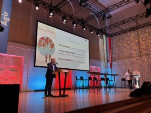 Intervention d’ouverture du symposium sur le harcèlement à l’Arsenal de Metz, avec un intervenant au pupitre devant un écran présentant les noms des personnalités d’ouverture.