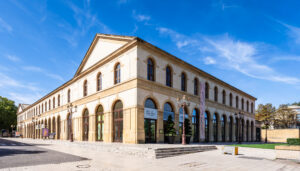 Vue d’angle de l’Arsenal de Metz sous un ciel bleu, montrant la façade en pierre, les arcades et l’entrée du bâtiment.